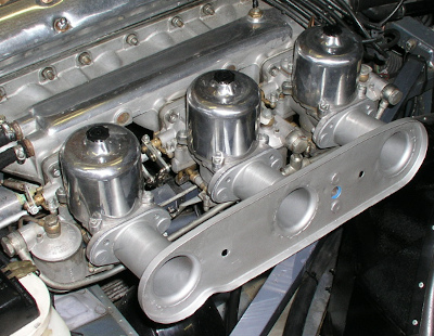 Three SU carbs lined up in the engine compartment of a 1960s era Jaguar E-type. The carbs look like polished aluminum bell-shaped cylinders.