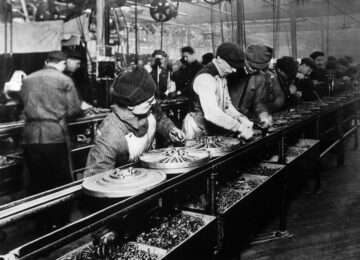 A line of about half-dozen men, bundled up against cold, affix bolts and assemble magnetos for Ford's Model T. The image shows the first assembly line created for Ford's car assembly line in 1913.