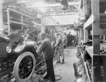 Working men lean over line of half-assembled Ford Model Ts in a factory in Copenhagen, Denmark. The black-and-white photograph depicts a line of men in essentially the same pose, showing the nearest man with is arms extended into the car's interior. Other men are shown in like poses into the distance.