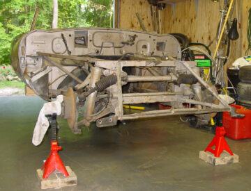 A stripped down chassis of a 1963 Jaguar E-type with badly worn light brown "sand" paint. The chassis has no engine and is stripped bare. It stands on jackstands.