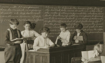 Grade school children surround a teacher's desk as she grade papers in a black-and-white photograph from about 1915. The students are in the lower grades, and they are standing in front of a blackboard covered with finely wrought handwriting.