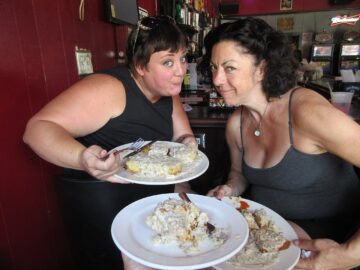Two women in black dresses lean toward each other as they show off three plates of biscuits and gravy. The plates are white and the gravy on the biscuits is white. The women are musician Megan Harris Brunious and singer Ingrid Lucia who are pictured at Buffa's Bar & Restaurant in New Orleans, Louisiana.