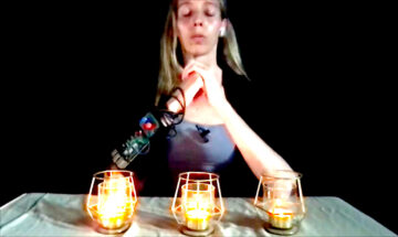A woman, out-of-focus against a dark background, clasps her hands. Her eyes are closed, as if in prayer or meditation. An electronic device is wrapped around her left forearm. Three lit votive candles burn in small clear-glass cup before her.