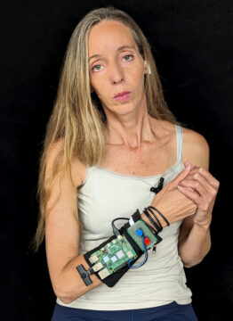 Avital Meshi, a performance artist who uses AI in her work, stands with herright arm raised to show an electronic device strapped on her right forearm. She has her head slightly cocked, her long blonde hair falling over her right shoulder. She is not smiling as she looks straight into the camera. The background is black, so her upper body is clearly outlined.