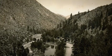 Black-and-white photo dating from mid-twentieth century (probably) showing the Poudre River and Colorado Highway 14 side-by-side winding through steep sanyon slopes. Next to the road in the middle of the picture are clustered dark buildings with sloped roofs. The cluster of buildings make up the “Mishawaka,” a resort and dance hall founded by Walter S. Thompson in the early 1910s.