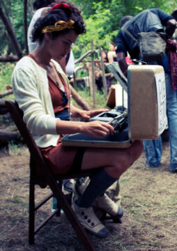 A woman in a maroon dress with a white sweater sits on a wooden folding chair. She is typing and looking very intently at the vintage typewriter in front of her. The typewriter is in a case, and the case is open. Tacked to the front of the case is a sign. It is illegible, but it says "POEM STORE."